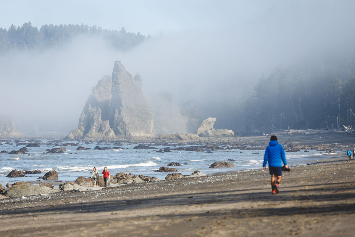 Garrett in a bright blue jacket and holding a camera walking along Rialto Beach on a foggy day with a view of giant rocks and trees.