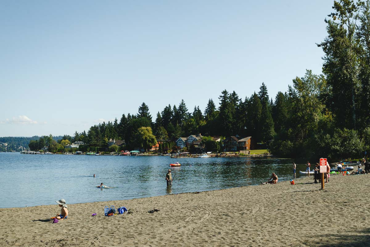 A few tourists hanging out on a beach  of Lake Sammamish in Idywood Park with lake houses in the background.