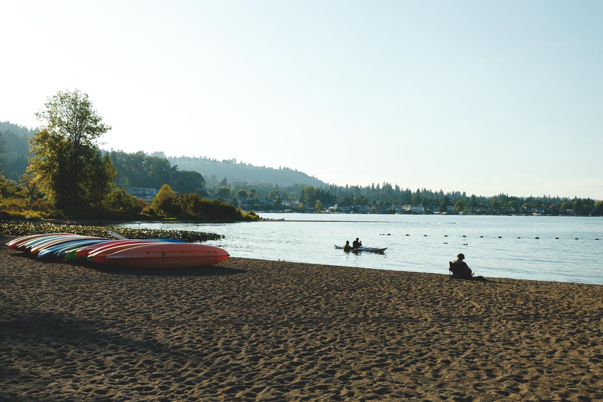 Two people taking a kayak out in the waters of Lake Sammamish while another person sits on the beach besides a line of kayaks.