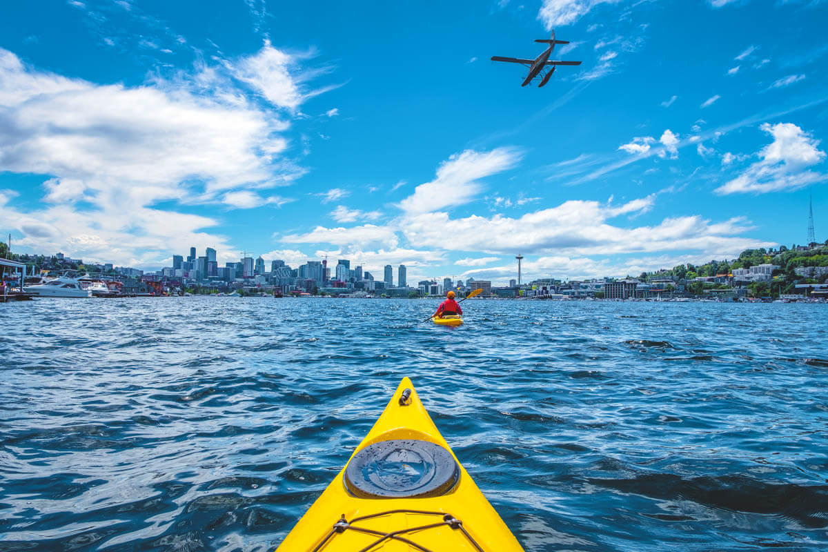 Two people kayaking on Lake Union with a view of downtown Seattle and a seaplane flying overhead.