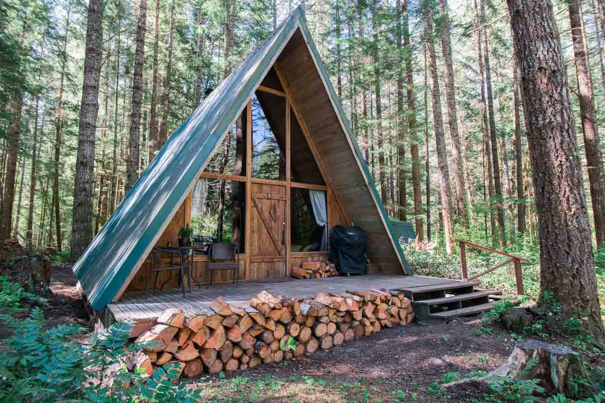 An A-frame cabin in the middle of the Washington woods with piles of fire wood out front.