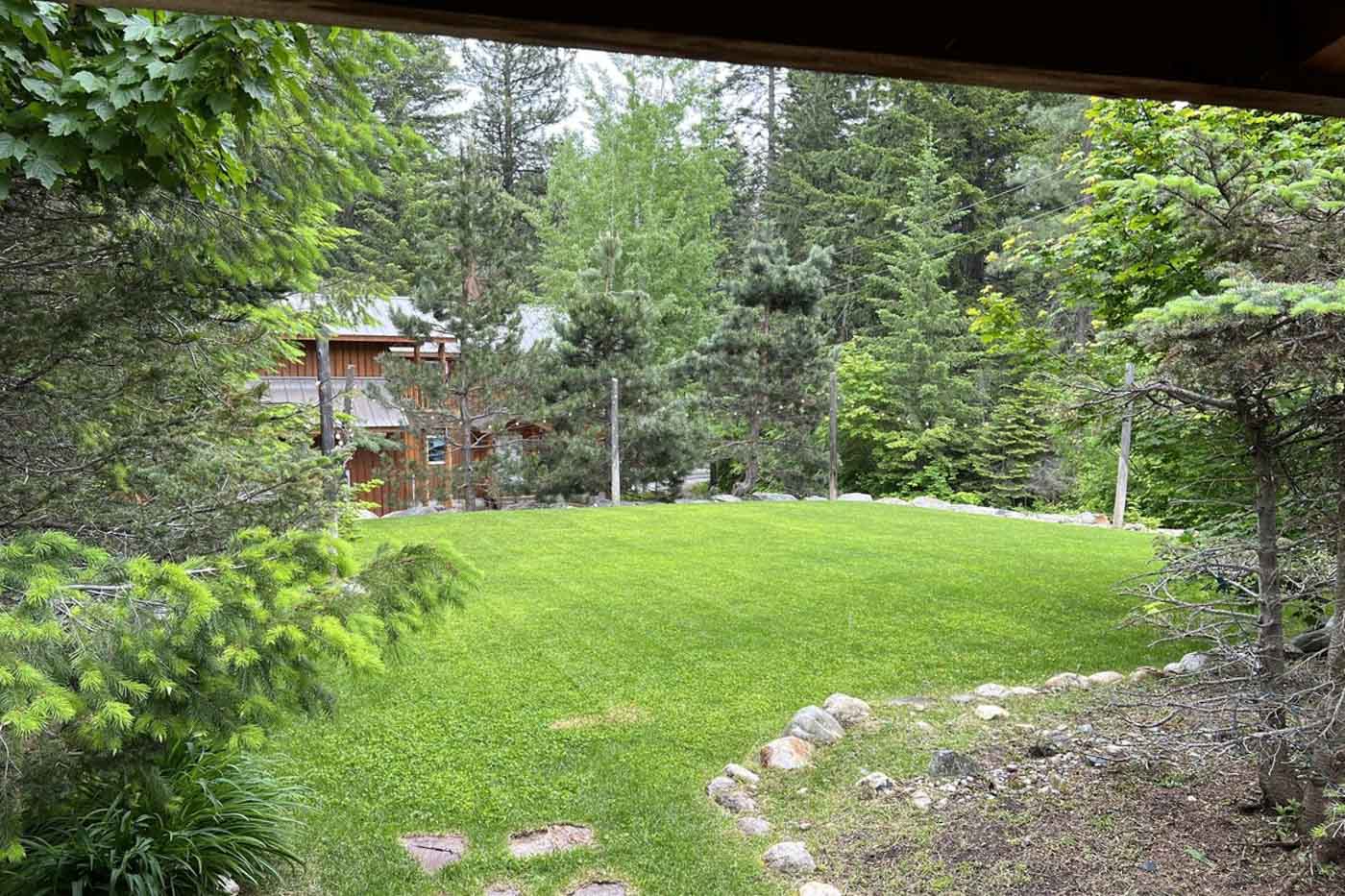 A view over a well groomed back yard surrounded by trees and a wooden cabin in the distance.