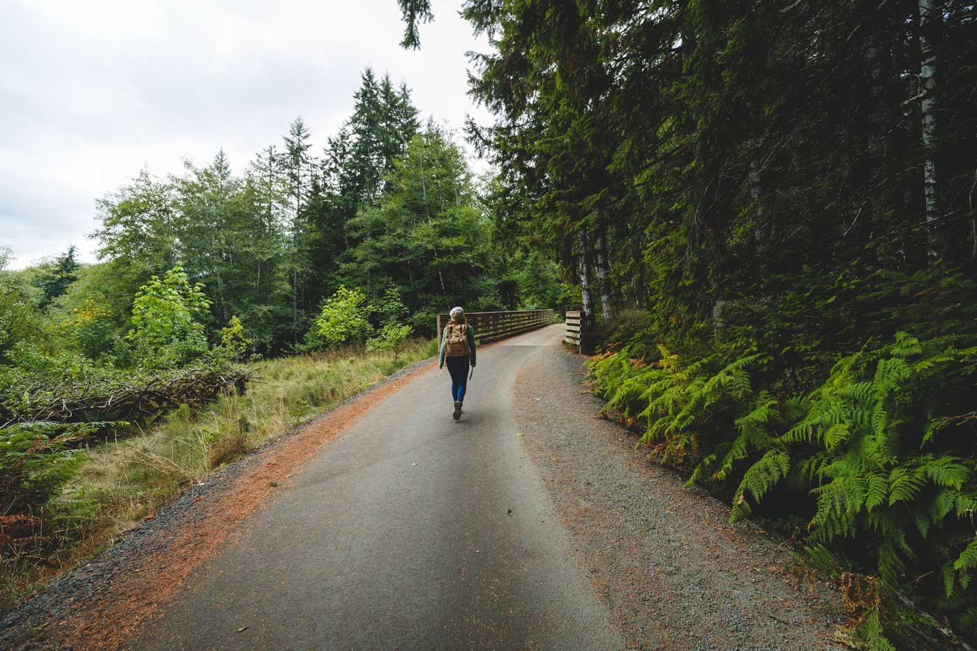 Nina in hiking gear and a backpack walking along a road towards a wooden bridge surrounded by trees along the Spruce Railroad Trail.